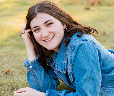 A senior girl lays on her stomach in the grass at Shannon Springs Park in Chickasha Oklahoma for her senior photo session. She smiles at the camera.
