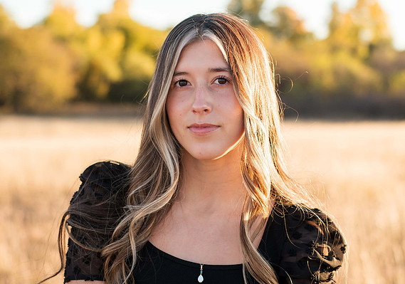 pretty senior girl with long blond hair in a black shirt looks softly at camera with sunlight hitting her hair and fall trees behind her standing in a field in Tuttle, Oklahoma