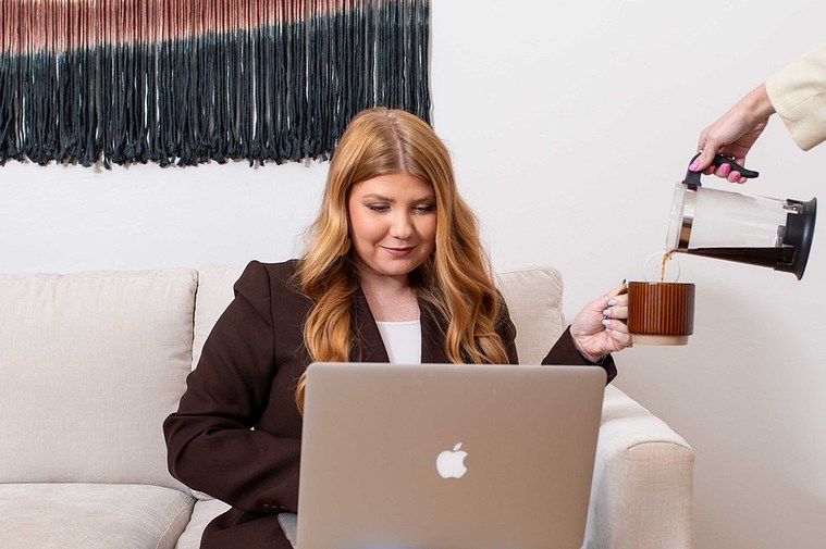 branding photo of a young female business owner sits working at her laptop on the couch while her business partner's hand is pouring coffee into her cup from a french press 