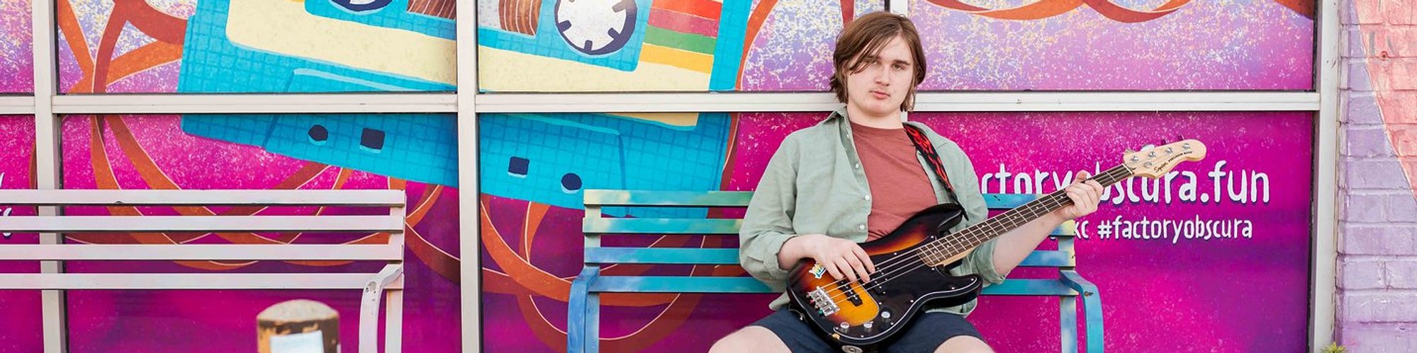 senior boy sitting on a bench in front of a bright mural wall holding a bass guitar in midtown Oklahoma City