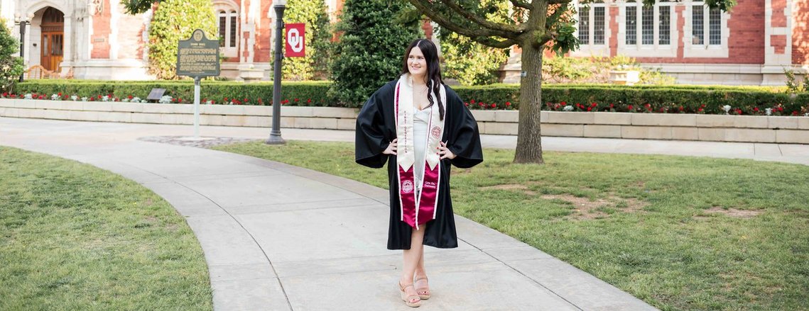 OU senior girl stands on a sidewalk on campus grounds wearing her graduation gown in Norman, Oklahoma