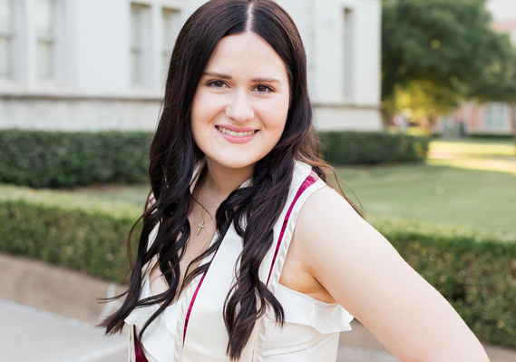 OU college senior girl with long dark hair wearing a white dress and graduation stole smiles standing in front of the chemistry building on OU campus in Norman, Oklahoma
