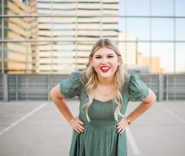 high school senior girl with blond hair and pink lipstick wearing a green dress has her hands on hips and smiles with the Devon tower behind her on the parking garage rooftop