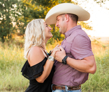 a couple in their 30s in western clothes holding hands, facing each other and smiling at one another in a field in rural Oklahoma