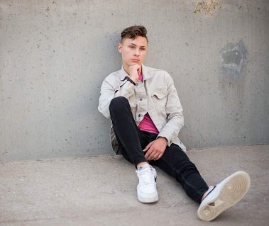 a teen boy sits against a parking garage rooftop wall with his chin in hand and one leg stretched out 