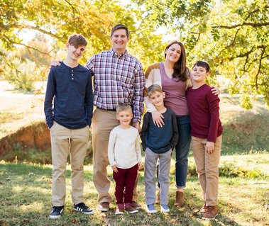 a mom, dad and their 4 boys stand and smile together in front of green trees at Will Rogers Gardens in Oklahoma City