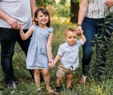 a little girl and toddler boy stand between their parents holding their hands and smiling