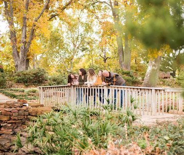 a family of 5 stand together on a foot bridge and look at one another surrounded by fall foliage at Will Rogers Gardens in Oklahoma City