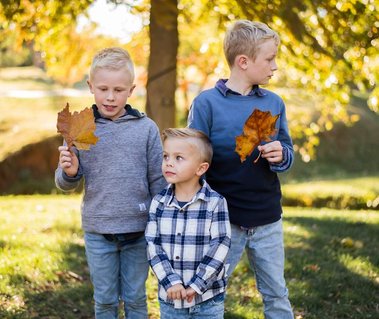 3 young brothers stand together holding leaves and looking away in front of trees at Will Rogers Gardens in Oklahoma City