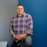 Man with glasses sitting in studio with colorful shirt.  Clothing styling by Image Concepts.