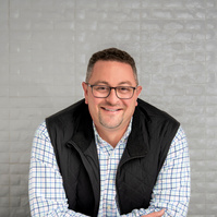 Man with glasses leaning on table with plans in vest jacket. Clothing styling by Image Concepts.