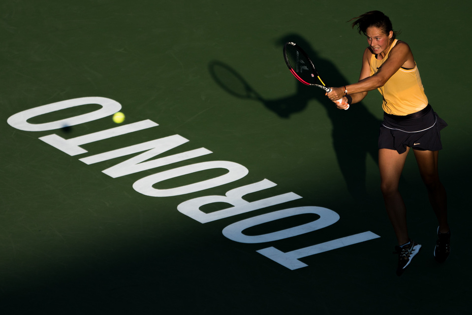 A female tennis player returns serve in the shadows next to a Toronto sign as the sun sets.