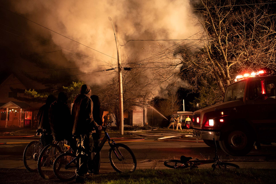 A group of people watch a house fire being extinguished by a fire department.