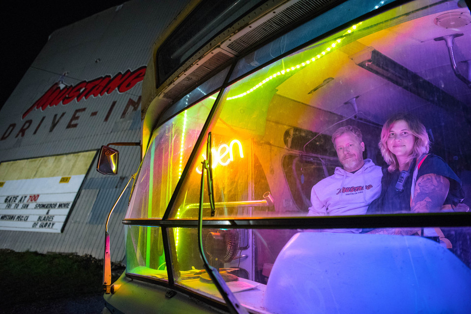 Drive-In theatre owners sit in a bus, that serves as their box office, with colourful lighting.