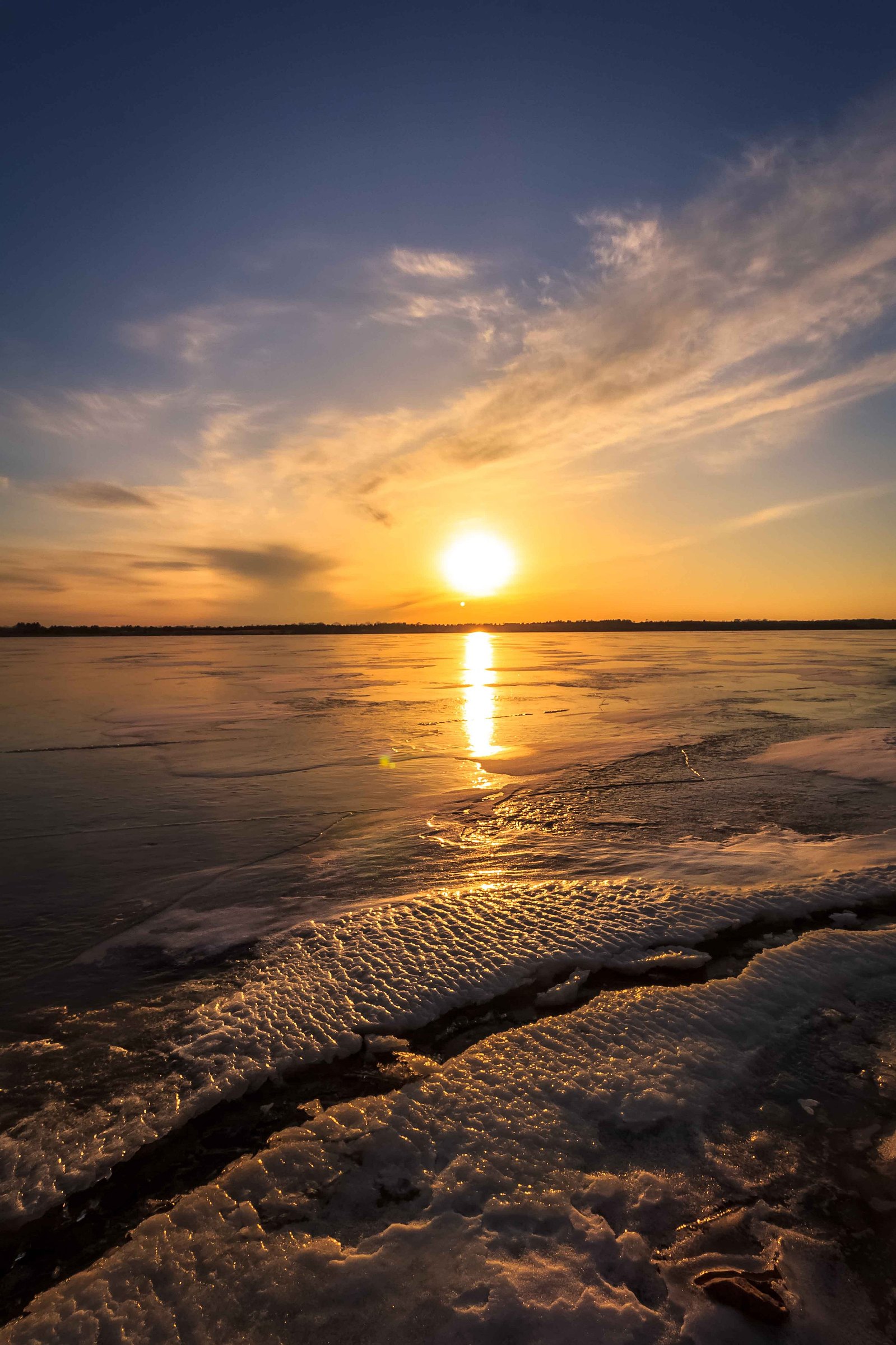 A dramatic sun lights up this image of a frozen lake with cracked ice in the foreground.