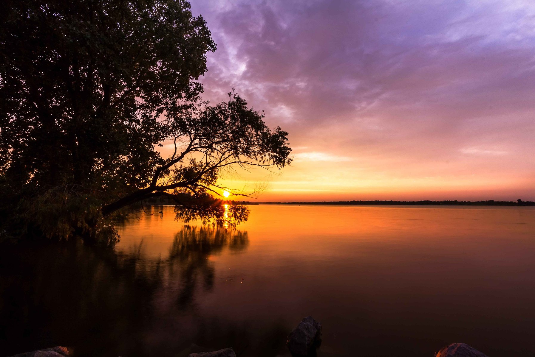A dramatic sunset sky filled with purples, pinks, orange and blue over a still lake against a silhouetted backdrop in Nebraska