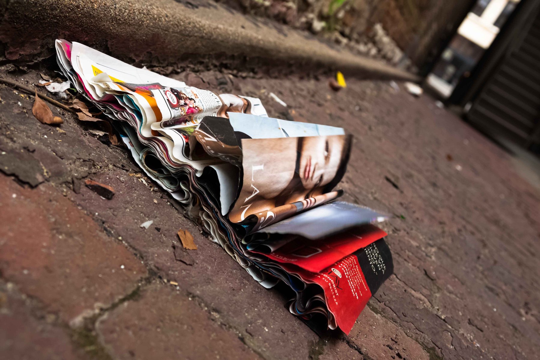 A wrinkled and torn magazine with a woman's face on it rests in a dirty alley.