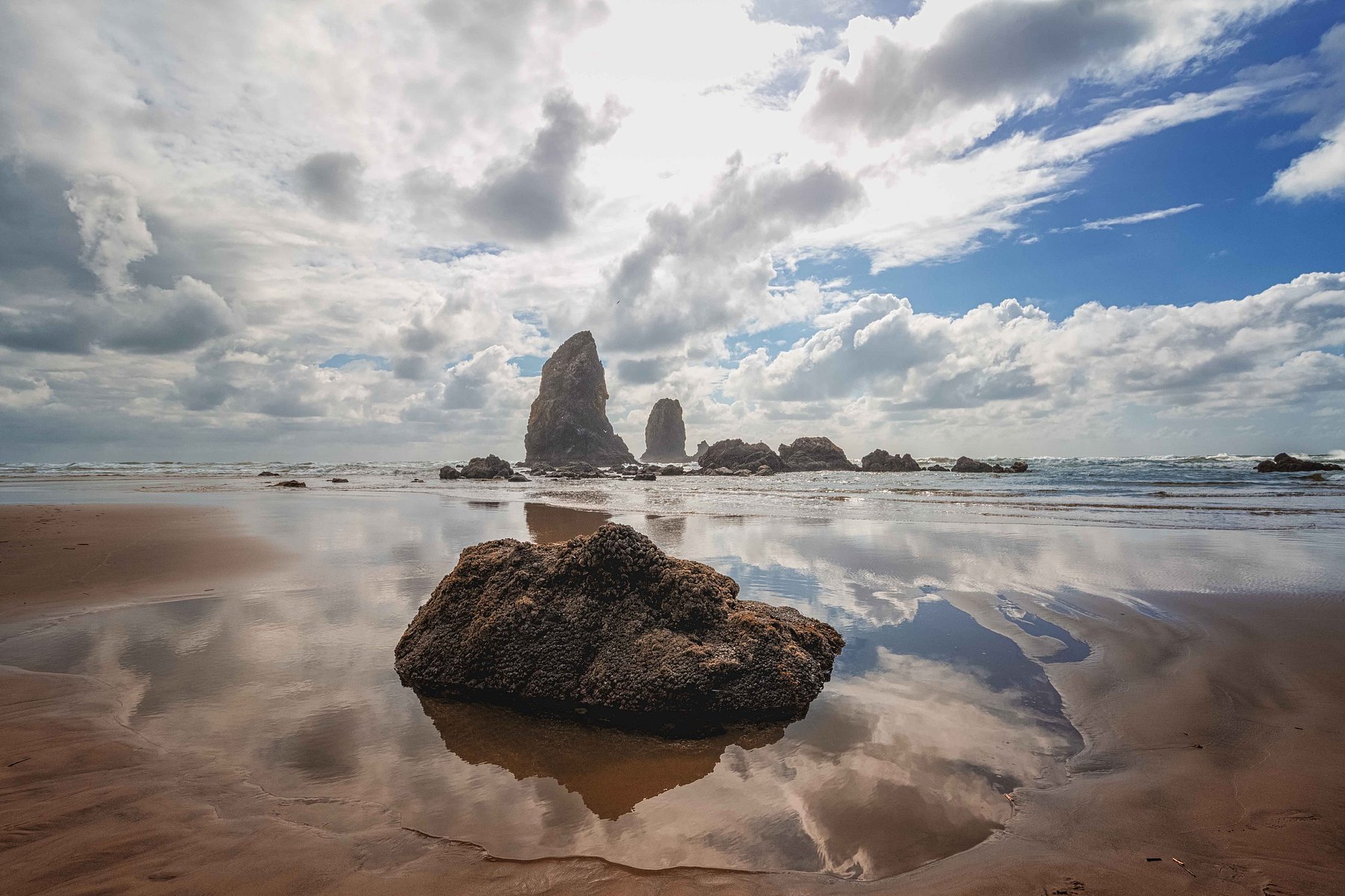 Standing water reflects swirling clouds on the beach with rocks jutting out of the foreground and in the far distance at Cannon Beach in Portland Oregon.