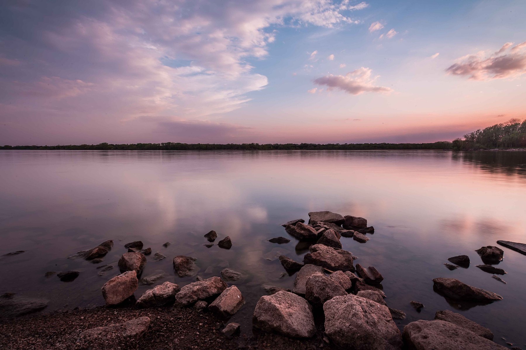 Rocks just out of calm smooth pink and purple waters that perfectly reflect a gently cloudy sky.