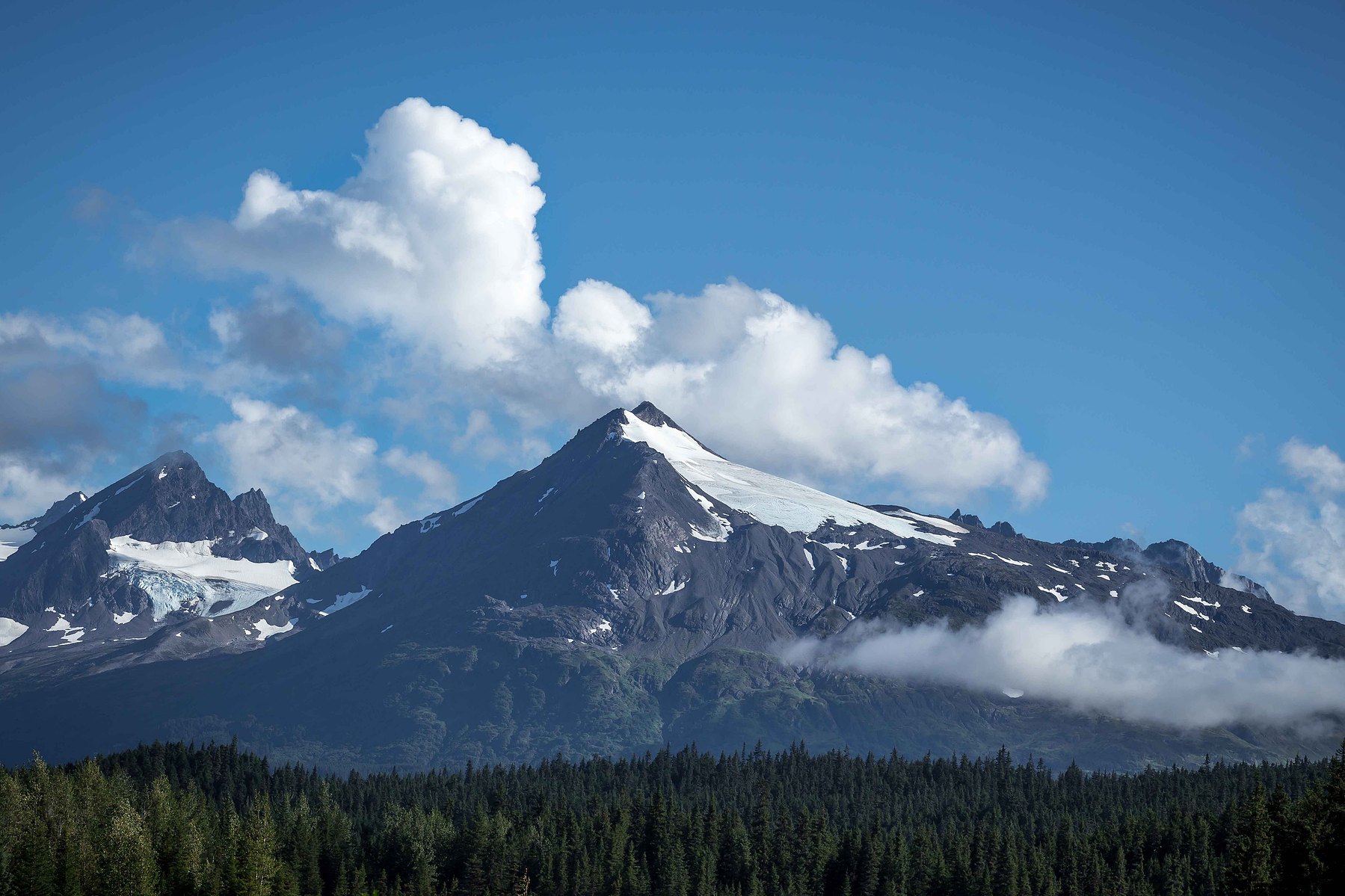 Clouds rise above majestic mountains against a clear blue sky in Alaska.