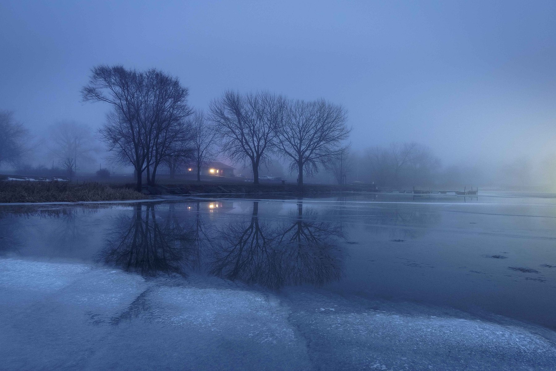 A cold chilly January morning at a frozen lake with leafless trees surrounding a house with a single light on against a icy blue sky at Pawnee Lake in Nebraska.