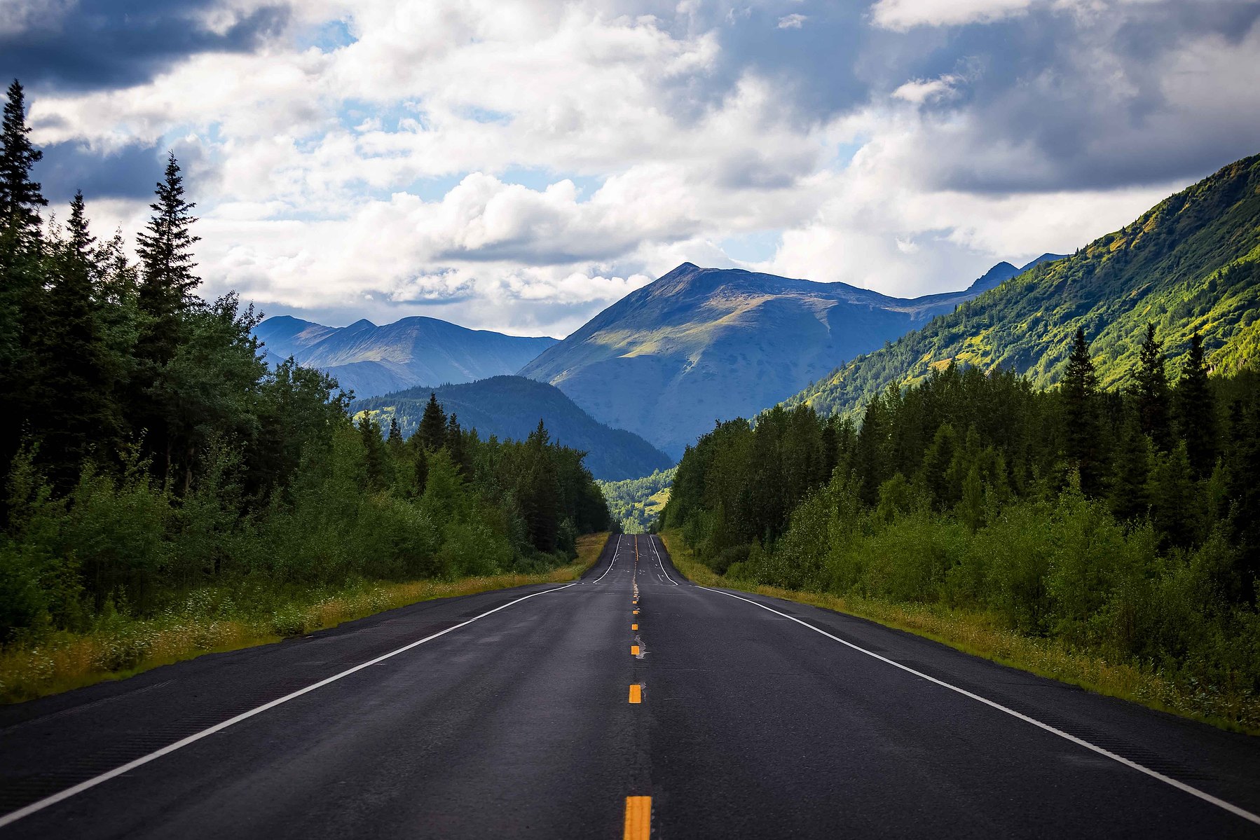 A concrete yellow striped road leads to majestic green mountains and cloudy skies in the distance in Seward Alaska.