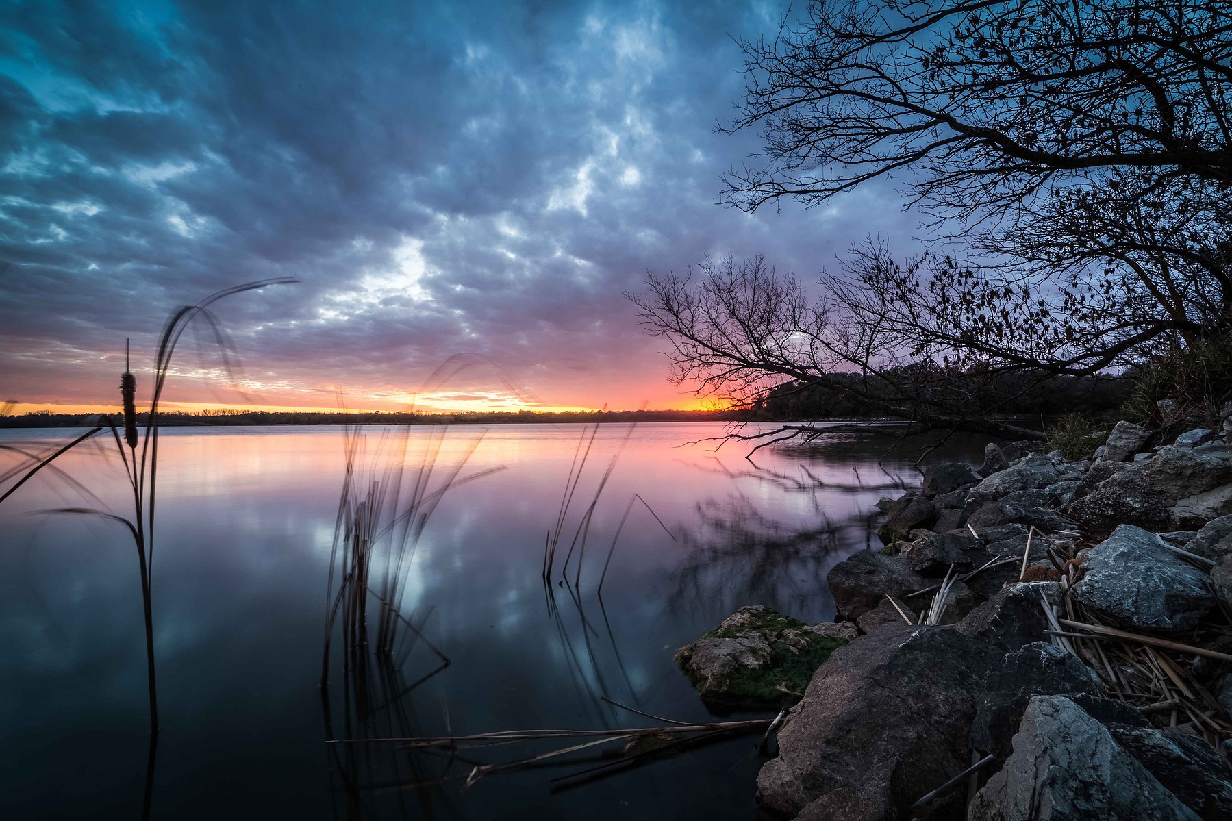 A dramatic sunset sky filled with purples, pinks, orange and blue over a still lake against a silhouetted backdrop in Nebraska