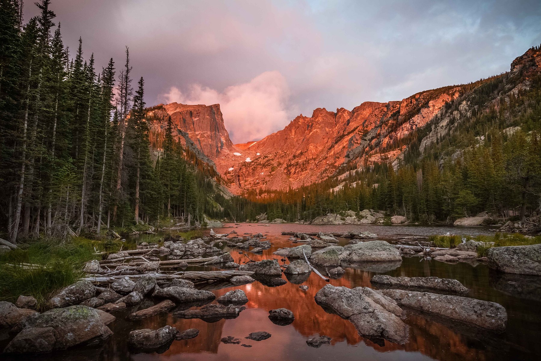 A still lake leading to a sun lit mountain in the distance in a forest in Colorado.