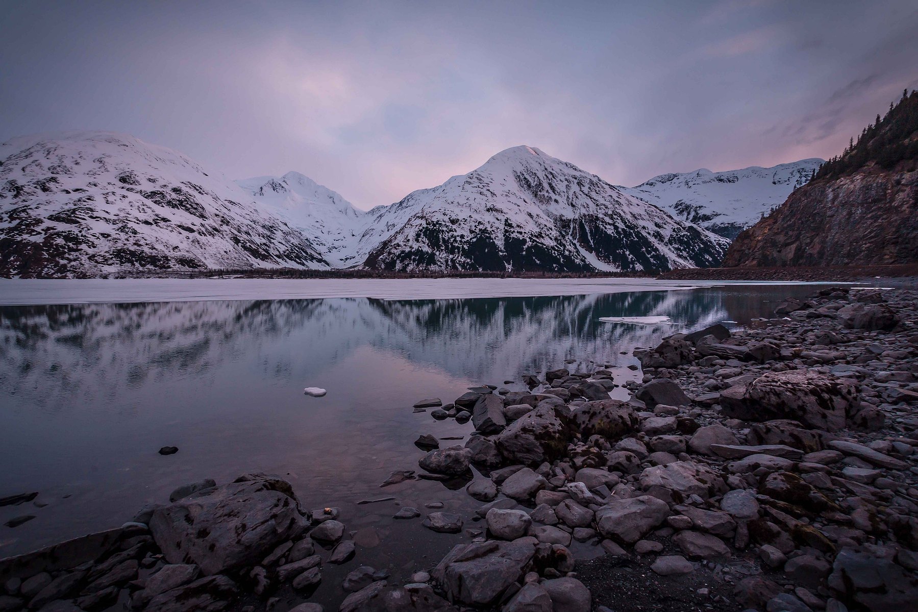The sun has all but set on the frozen mountains and rocks of the still lake at Portage Glacier in Alaska.