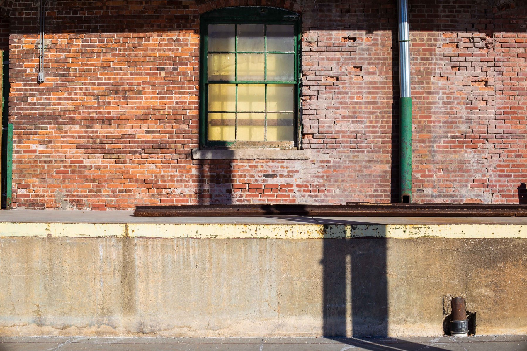 Shadows bend around a ledge leading to a solitary window in the side of an old brick building.