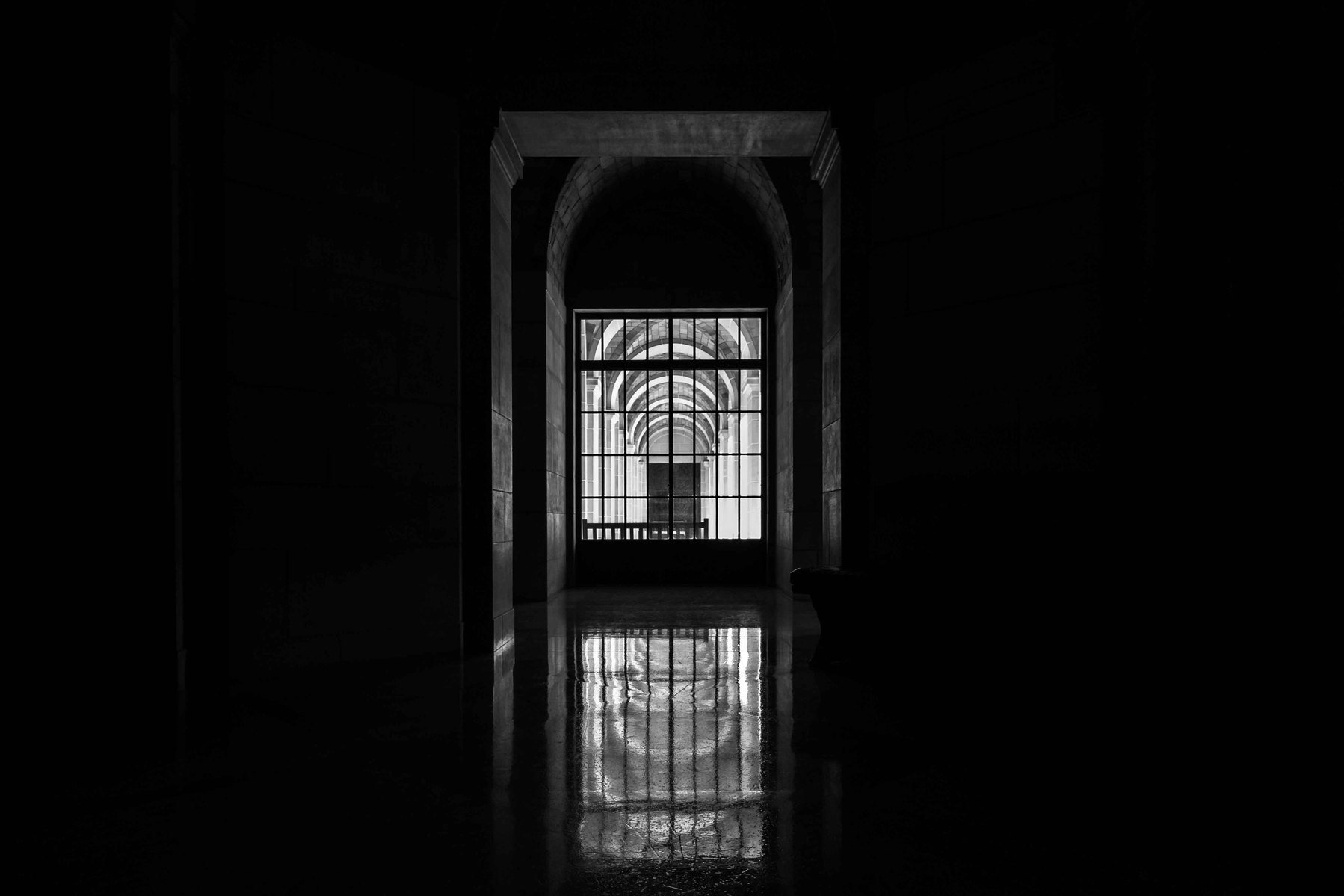 A pitch black hallway with a solitary light coming from a door at the end of it.