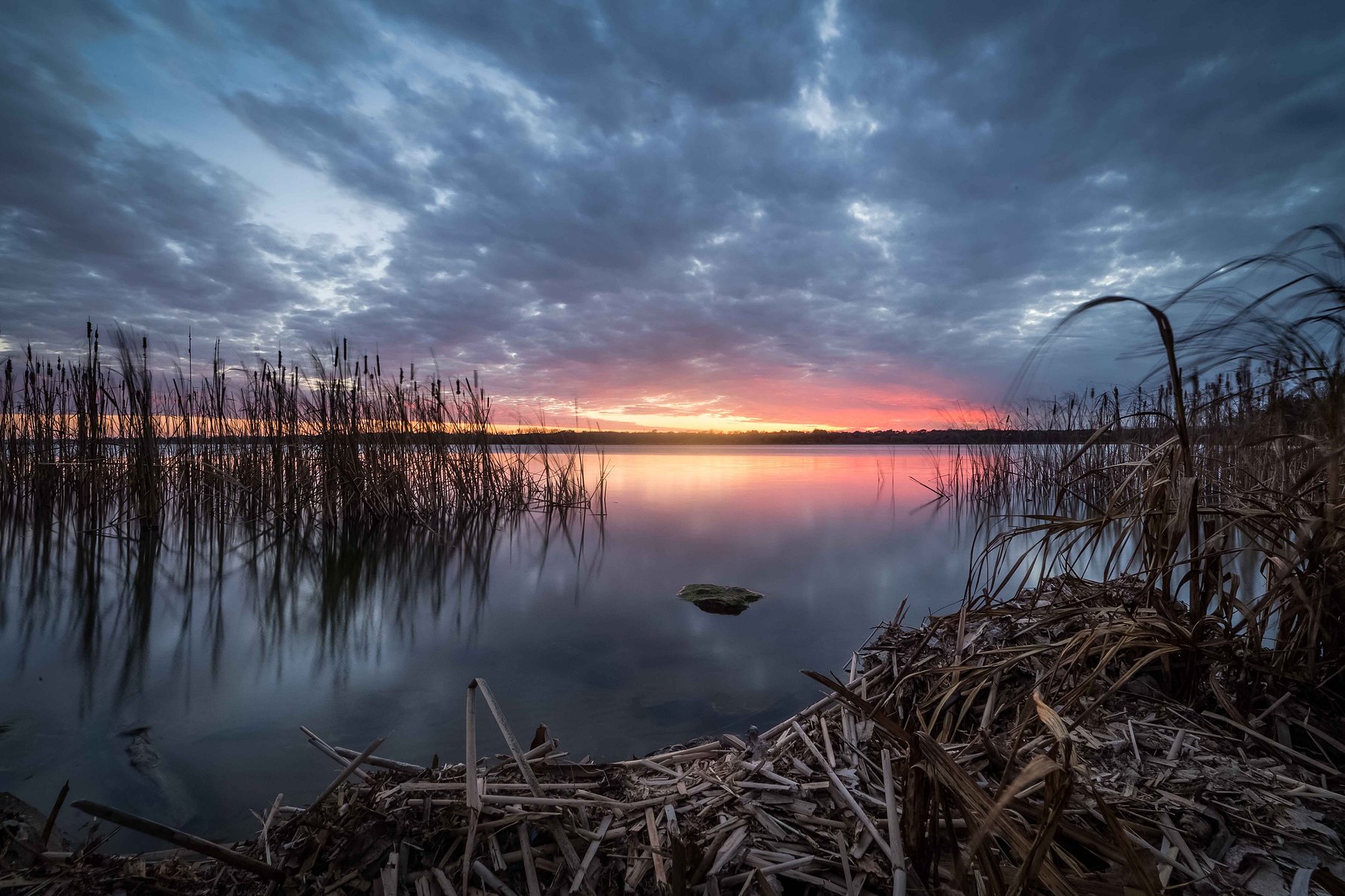 A dramatic sunset sky filled with purples, pinks, orange and blue over a still lake against a silhouetted backdrop in Nebraska