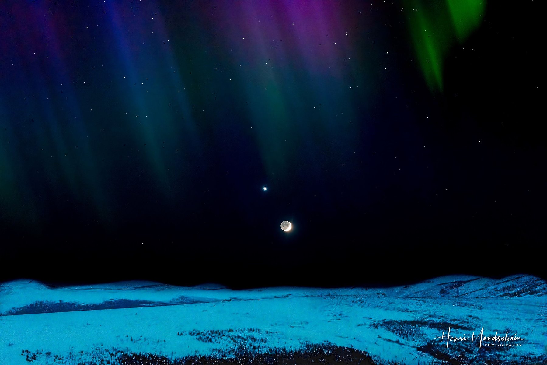 Colorful aurora curtains hang over the moon and Venus on a snow-covered landscape.
