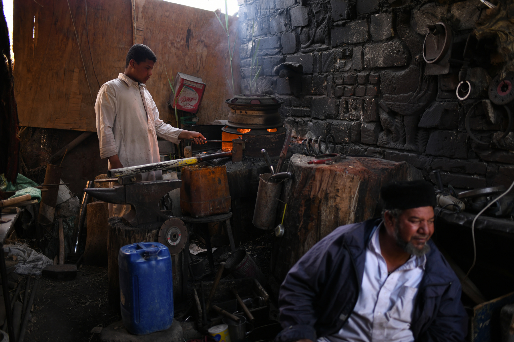 Omar Khattab’s fifteen-year-old son Badr fills a blacksmithing forge with coal while his father greets a guest at his workshop. Khattab says he has been teaching blacksmithing to his eldest son from an early age.
