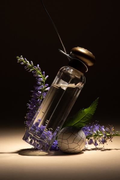 Flacon de parfum suspendue par un fil de métal avec des fleurs mauves, sur fond beige et noir. 