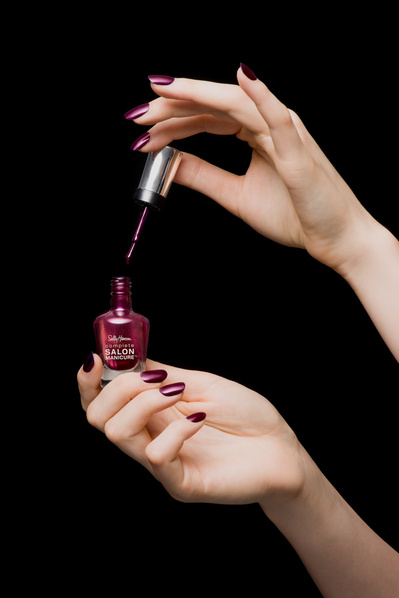 Mains de femme tenant un vernis à ongles violet ouvert, sur fond noir.