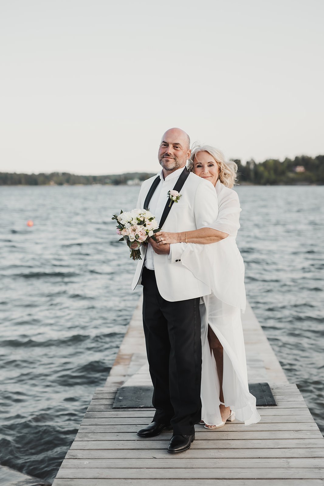 En kvinna håller om en man medan hon står bakom honom. Han klädd i vit kavaj, svarta byxor, hon i vit lång klänning och brudbukett i handen. De står på en trä brygga  till havs i horisonten syns fastlandet.
