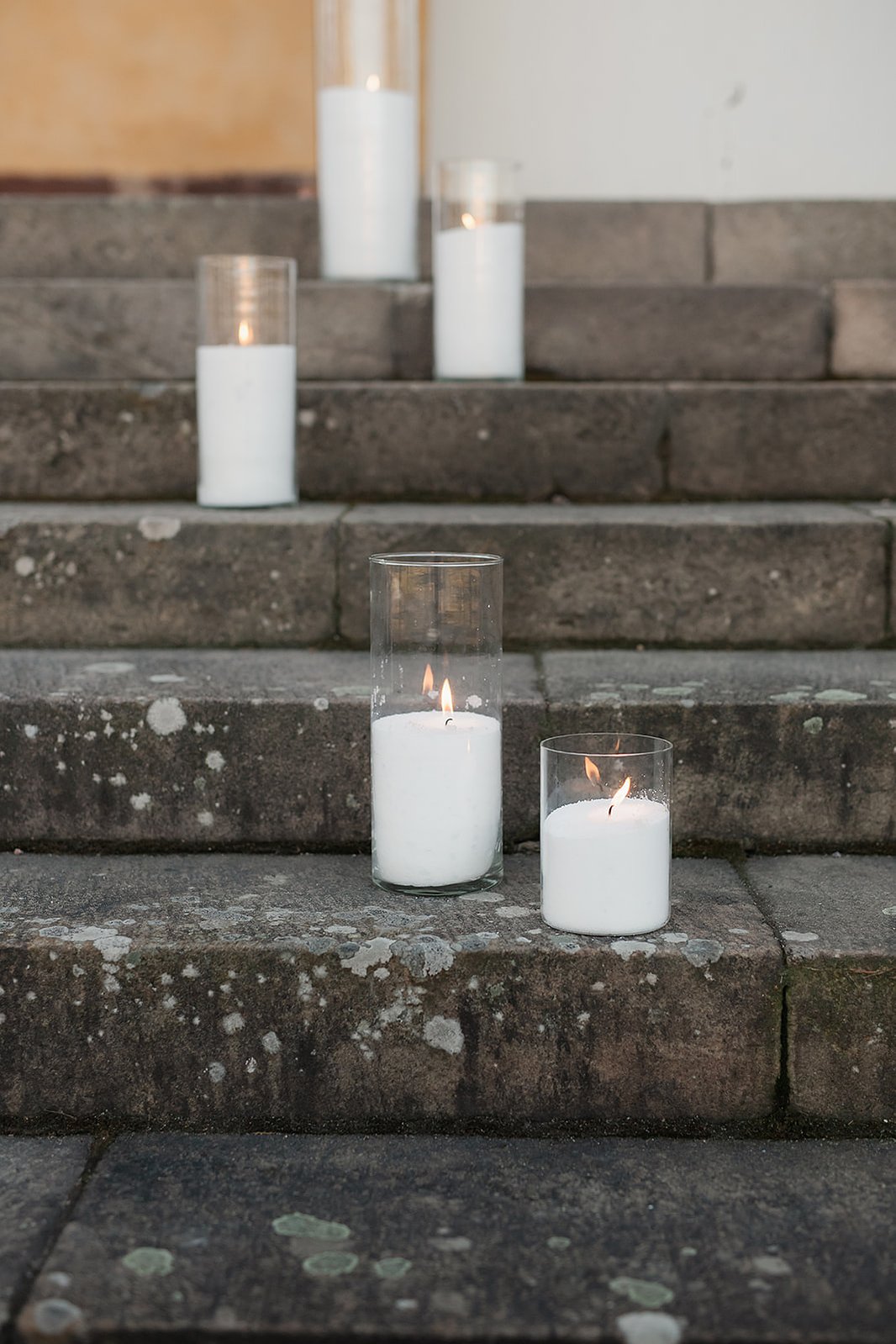 Fem tända vita ljus i glas cylindrar utomhus placerade på en mörk trappa utomhus.
