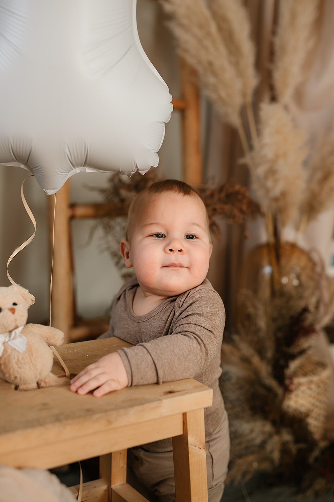 Liten pojke i beige tröja står vid en träpall med ballong och nalle inomhus.