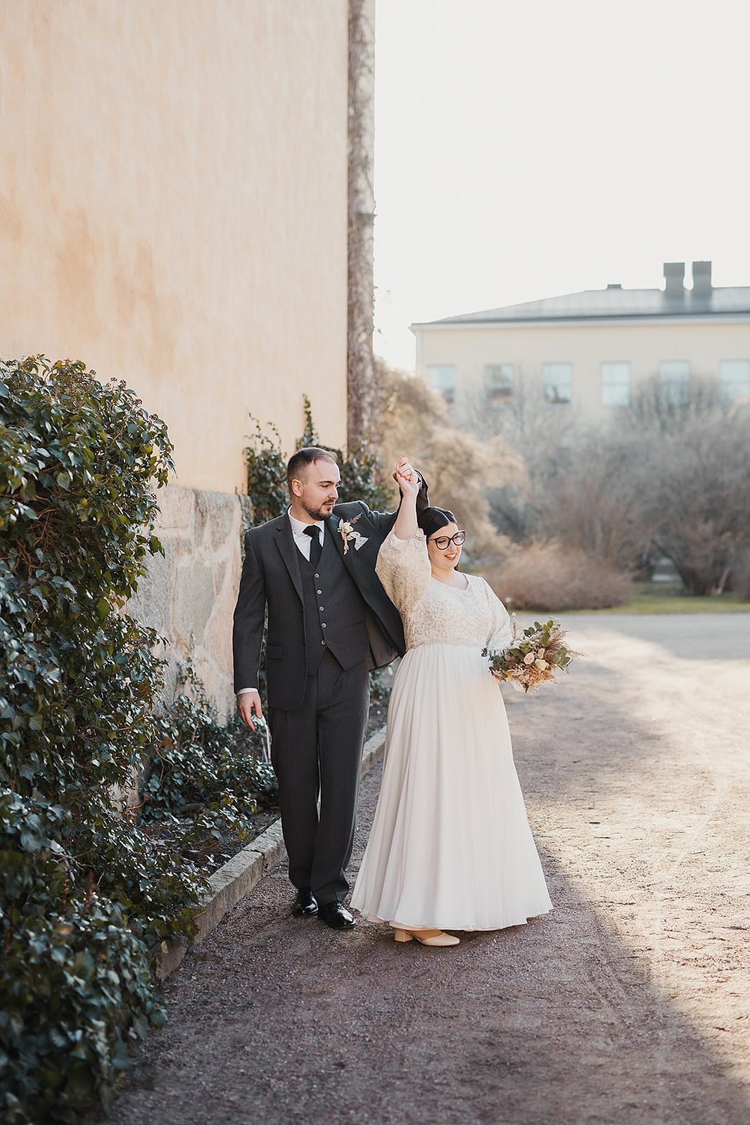 Ett par dansar utomhus i en trädgård med växter vid en gul fasad.  Mannnen i mörk grön kostym håller kvinnans hand. Hon klädd i vit lång klänning med blommor i handen.