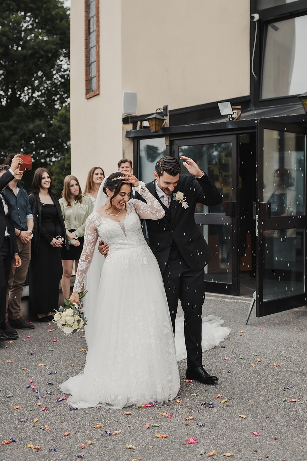 Kvinna i lång vit brudklänning står tätt ihop med en man i mörk kostym och blomma på kavajen. De håller vänster handen höjt mot ansiktet för att skyddas sig mot risgryn och godis regn utanför en kyrka. Bakom dem gäster som tar bilder.