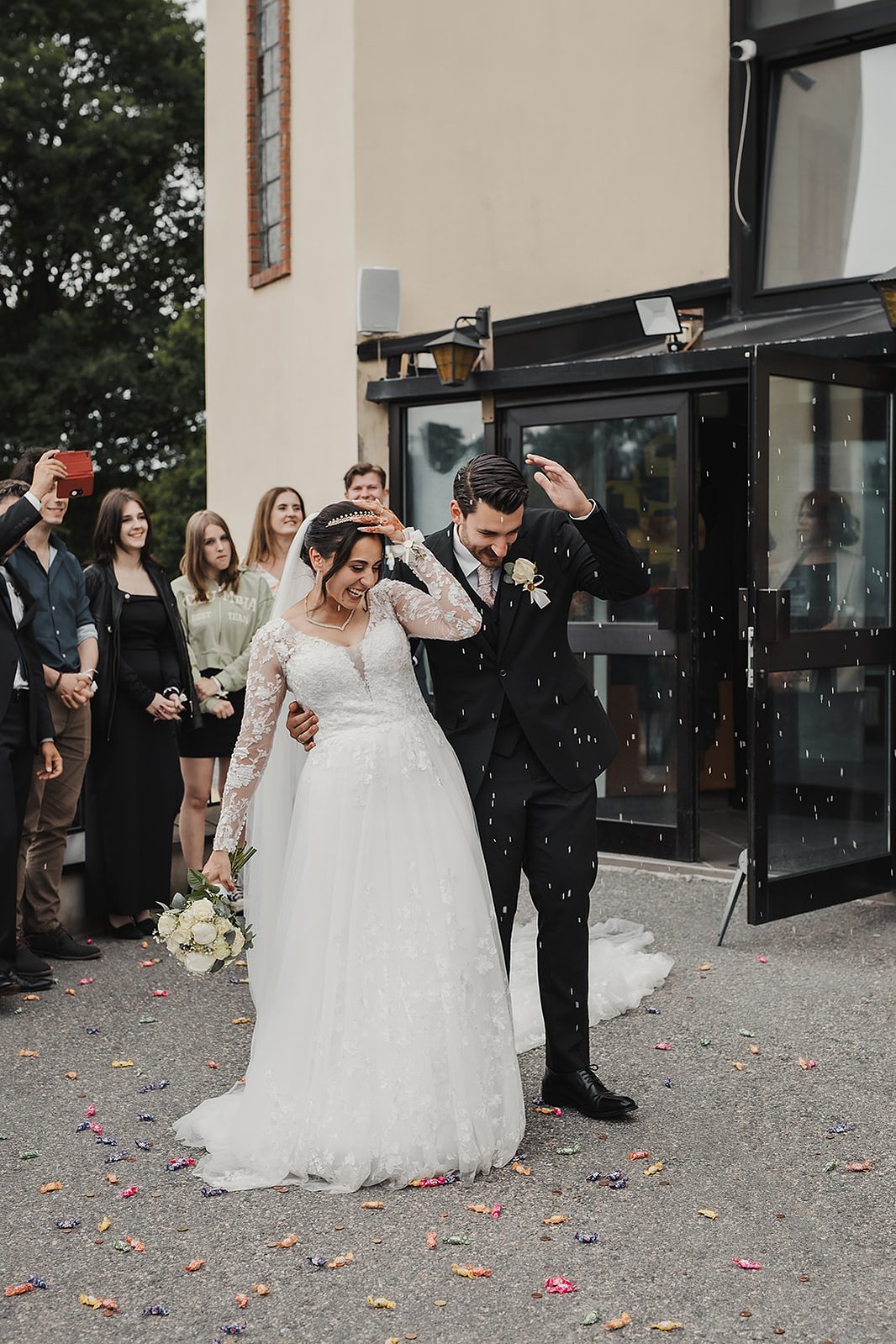 Kvinna i lång vit brudklänning står tätt ihop med en man i mörk kostym och blomma på kavajen. De håller vänster handen höjt mot ansiktet för att skyddas sig mot risgryn och godis regn utanför en kyrka. Bakom dem gäster som tar bilder.