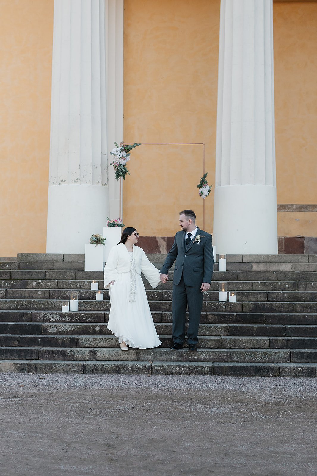Ett par går ner för en trappa och byggnad med flera vita pelare. Mannnen i mörk grön kostym håller kvinnans hand. Hon klädd i vit lång klänning med blommor i handen.
