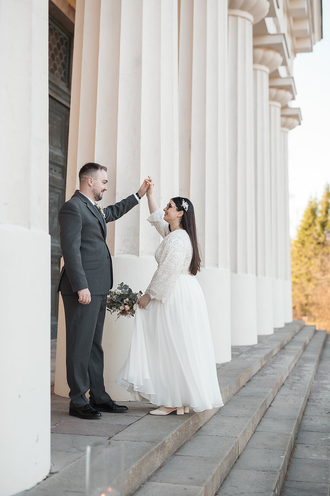 Ett par står mitt emot varandra vid en. trappa och byggnad med flera vita pelare. Mannnen i mörk grön kostym håller kvinnans hand. Hon klädd i vit lång klänning med blommor i handen.