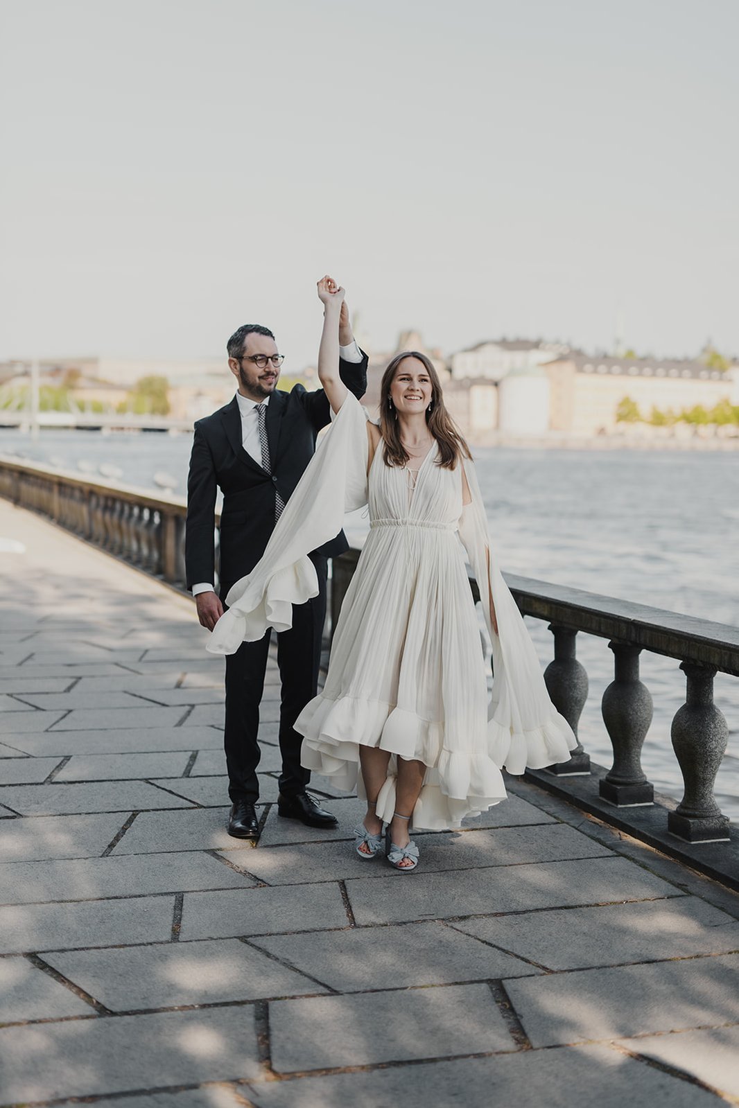 En man i mörk kavaj håller en kvinna i handen medan hon snurrar runt med sin benvita klänning och blå sommarskor, utomhus. I bakgrunden vatten från mälaren och husfasader från en ö.