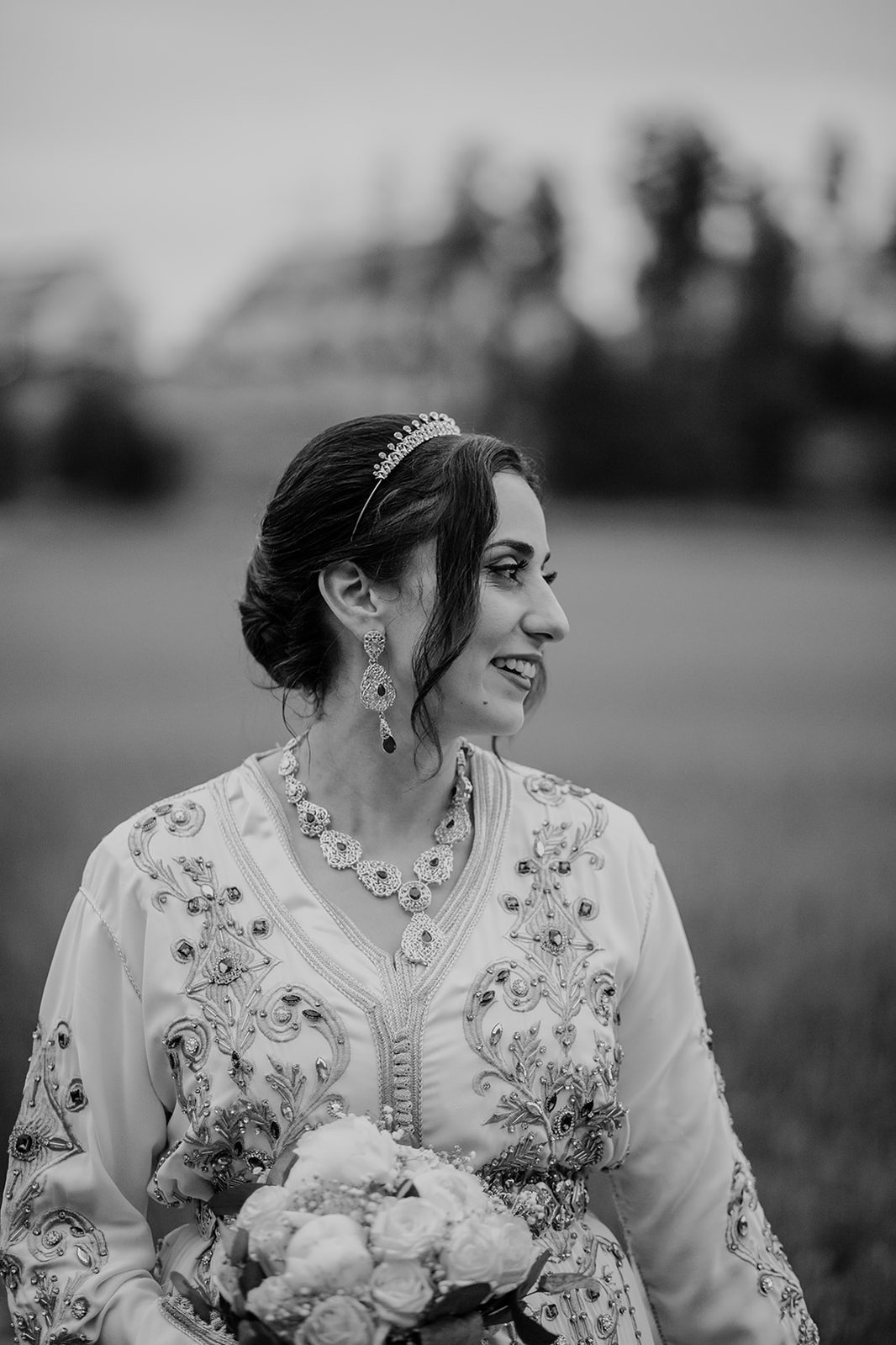Kvinna med uppsatt hår, diadem och långa guld örhängen och halsband i profil. Klädd i vit orient inspirerad klänning med stenar och håller i en bukett med blommor.