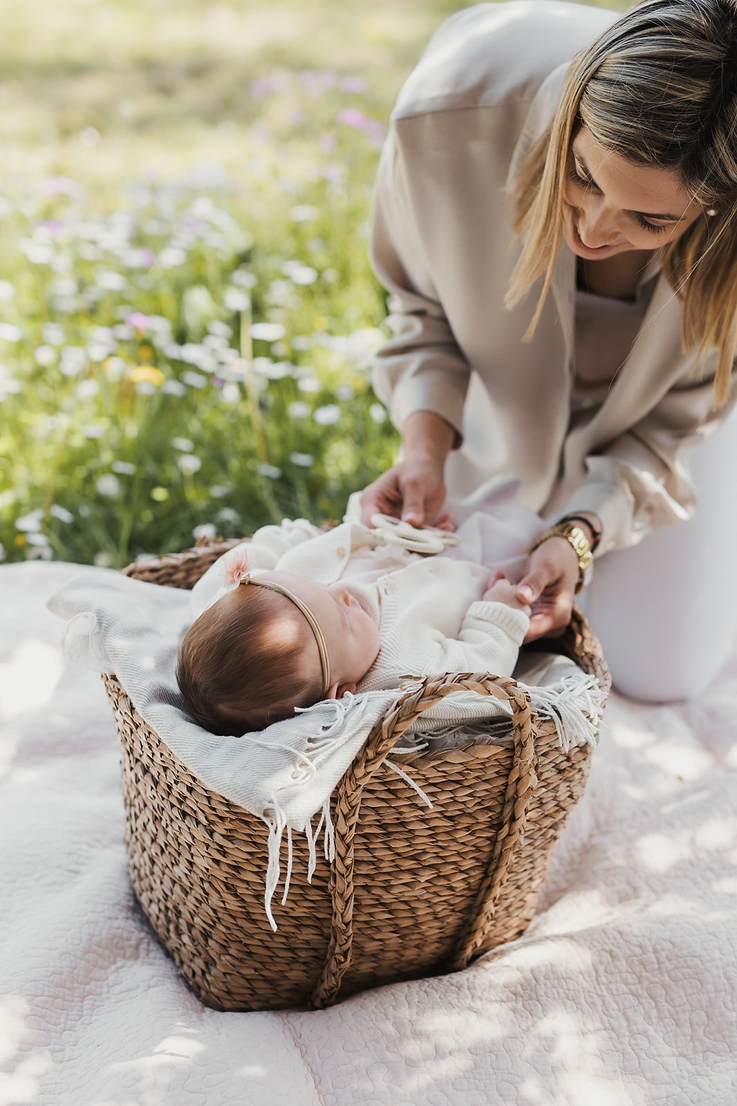 På en flätad babykorg och filt utomhus ligger en flick bebis medan hennes mamma lutar sig framåt.