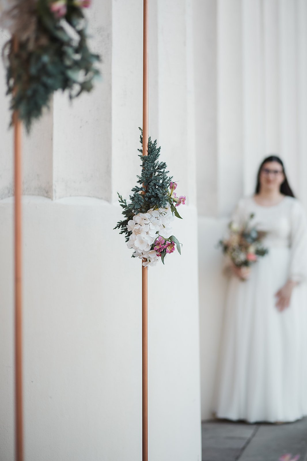 Vitt och grön blomdekoration på en kopparbåge. Bakom skymtar en kvinna med blommor i handen och vit lång klänning  bland vita pelare.