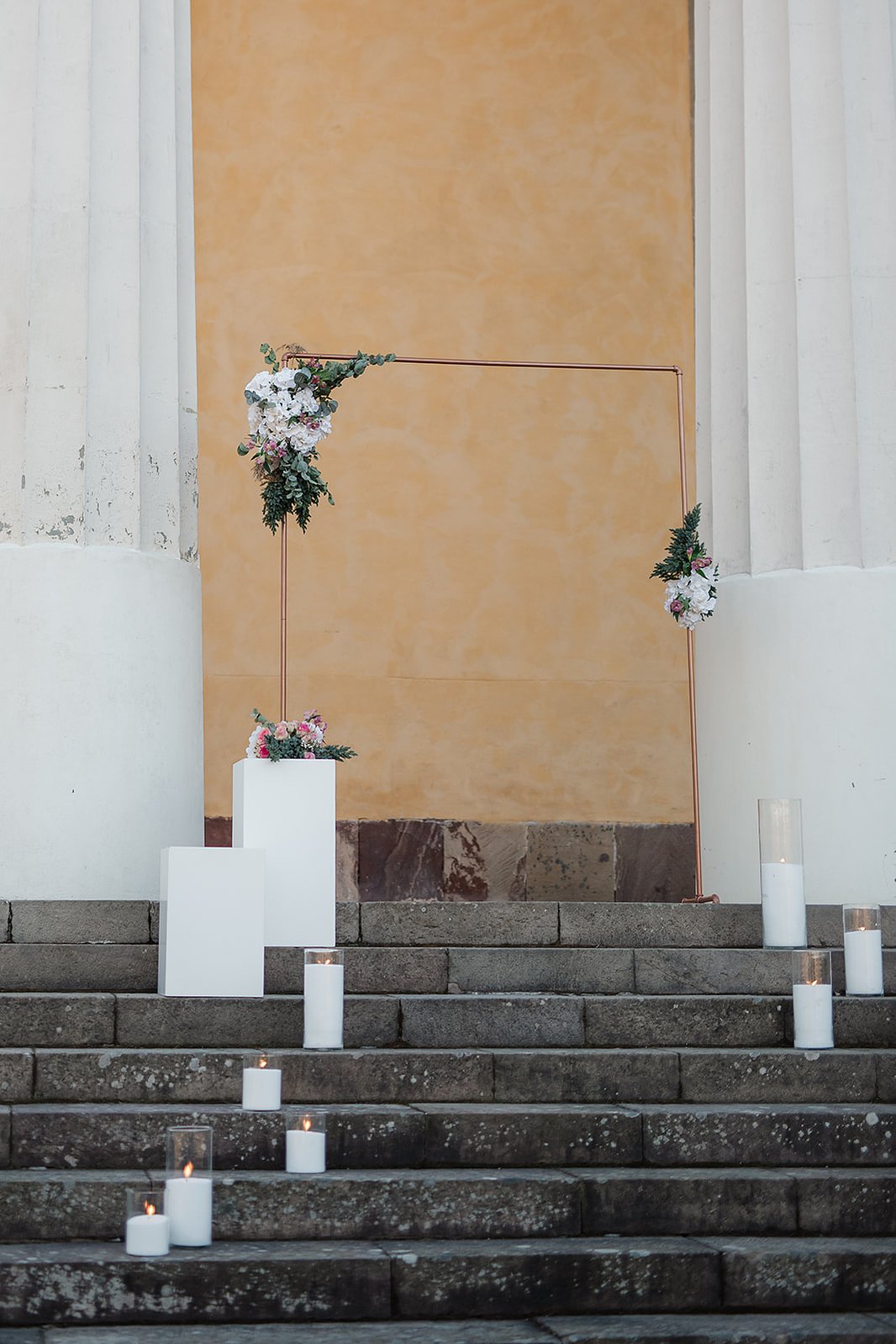 En mörk stentrappa utomhus med två vita pelare och gul vägg bakom. På trappan står en vigselbåge med blommor, två vita kuber med blommor och glas cylindrar med vita tända ljus.
