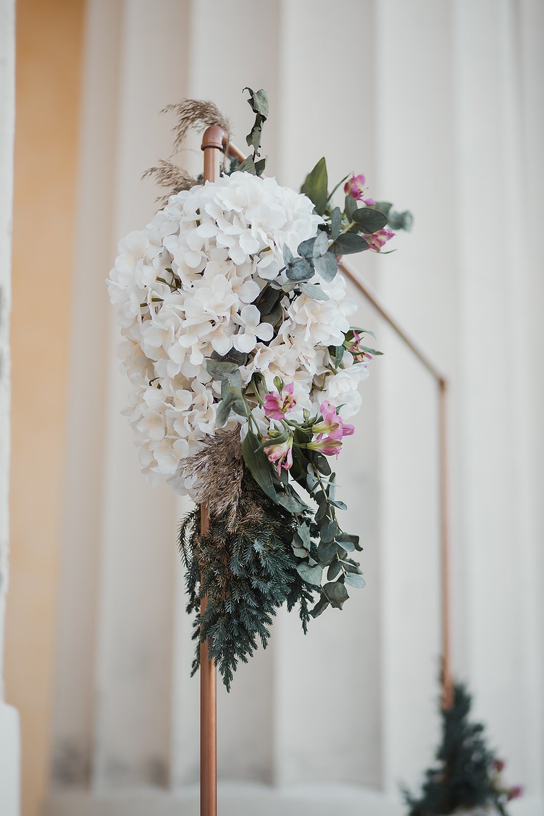 Vitt och grön blomdekoration på en kopparbåge. Bakom skymtar vita pelare.